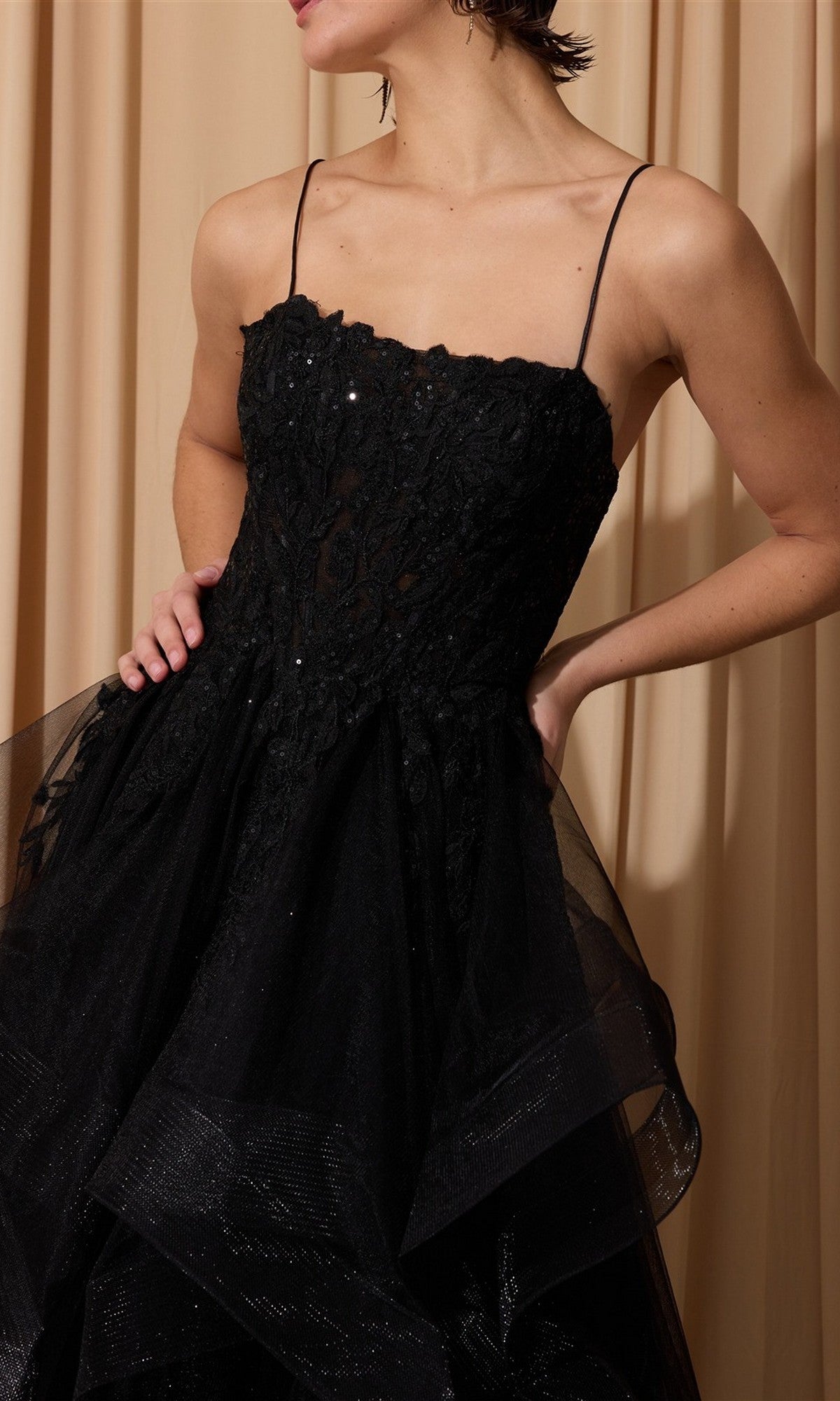 Wearing the Long Prom Dress: Eureka EK167, featuring a sleeveless black floral lace corset bodice with spaghetti straps and a layered tulle A-line skirt, standing before beige curtains.